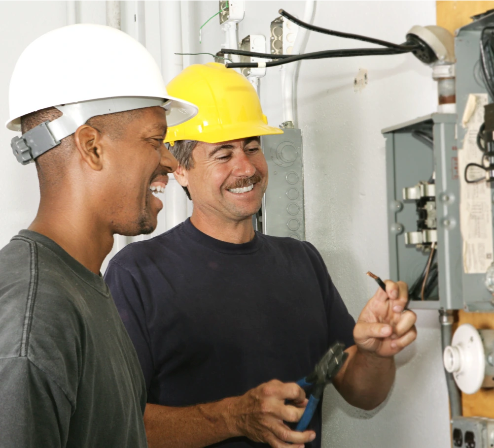 Two men perform work on an electrical box. One is holding pliers.